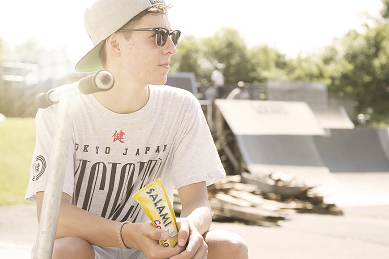 Jugendlicher mit Sonnenbrille und Cap sitzt im Skatepark und macht eine Pause. In der Hand hält er eine Packung Salamini. neben ihm steht sein Roller.